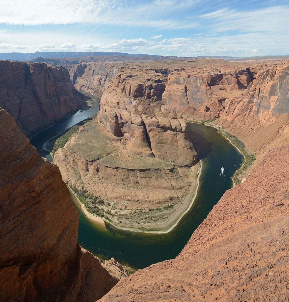 A green river curves in a tight bend around a large sandstone rock formation, surrounded by red canyon cliffs under a blue sky with thin clouds.